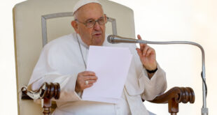 Pope Francis sits in a tall-backed white chair, holds a paper, speaks into a microphone and makes a pointing gesture