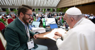A young man with brown hair wearing a suit smiles at Pope Francis as he signs a document